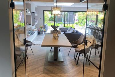 Wide view through a set of open, black steel-look double doors, looking from a herringbone floor hallway into a bright dining area