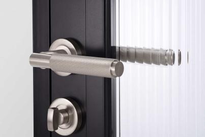 Close-up of a knurled stainless steel handle and thumb-turn lock on a black Origin OI-30 door with reeded glass