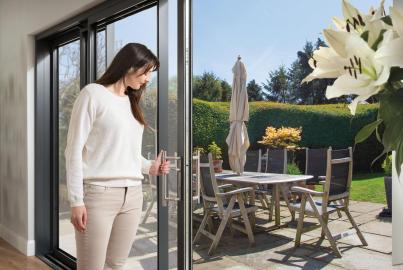 A woman slides open a dark grey Origin OS-44 sliding door, leading to a sunny patio with a table.