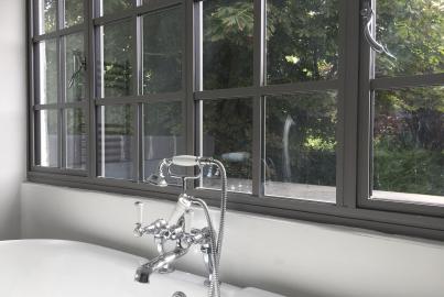 Close-up of a grey Aluco steel-look window with a grid pattern, set above a freestanding bathtub with classic chrome taps
