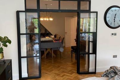 Fully open black Aluco steel-look double doors with a top panel, connecting a living room to a dining area