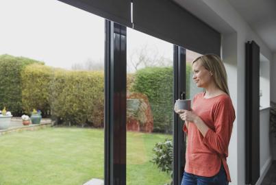 A woman holds a mug while looking out of a closed, dark grey Origin OB-49 bifold door onto her garde