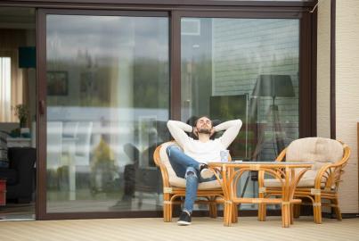 A man relaxes in a chair on a deck in front of a large, closed, dark-framed sliding door with reflections on the glass
