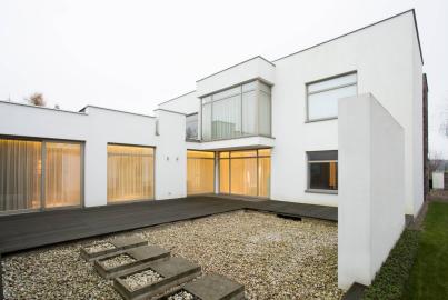 A modern, white minimalist house with large windows and a gravel courtyard, viewed on an overcast day