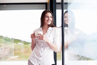 A woman smiles while holding a coffee cup, standing by an open Alitherm door and looking out