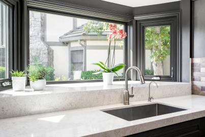 Modern kitchen sink with a large, dark grey corner window above the counter, showing an orchid and a view of a house