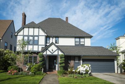 A two-story Tudor-style house with white stucco, dark trim, a grey roof, and a front lawn on a sunny day
