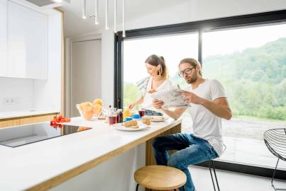 A couple eats breakfast at a kitchen island, in front of a large, black-framed sliding glass door with a hillside view