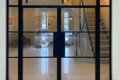 Closed black steel-look double doors with side panels, viewed from a hallway, looking into another hall with a staircase