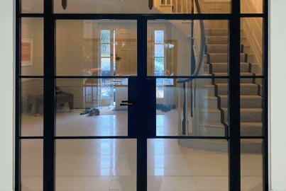 Closed black steel-look double doors with side panels, viewed from a hallway, looking into another hall with a staircase
