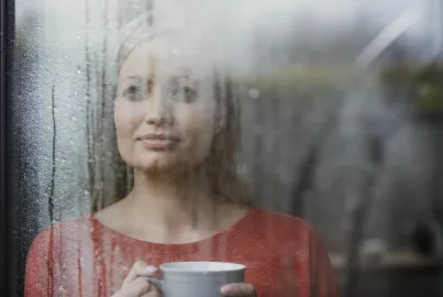 A person holds a mug and looks thoughtfully out of a large glass window that is wet with condensation
