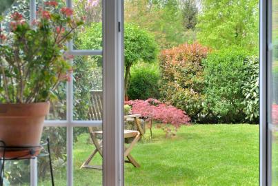 View through an open white Origin double door with grid panes, looking onto a lush green garden with a lawn and chairs