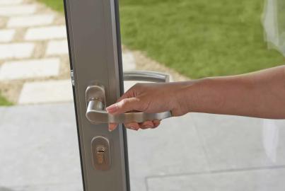 A hand turns the silver lever handle of an open grey Origin door, leading out to a garden patio