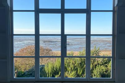 Interior view of a large white steel-look window, looking out over a sunny coastal landscape at low tide.